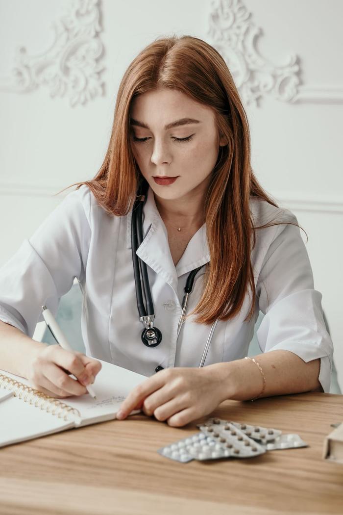 A healthcare worker in a white coat writing in a notebook at a desk.