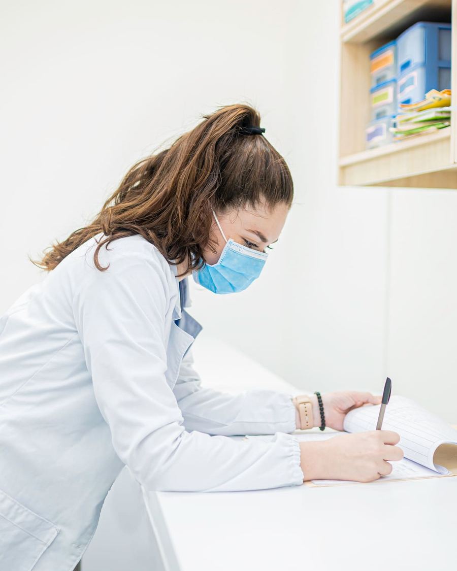A clinician in a white coat and mask writing notes in a logbook at a clinic counter.