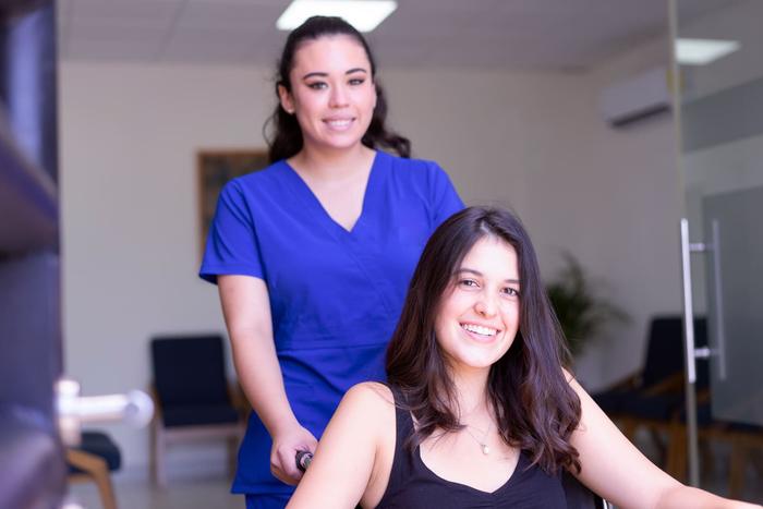 A nurse in blue scrubs helping a smiling patient in a wheelchair through a clinic waiting area.