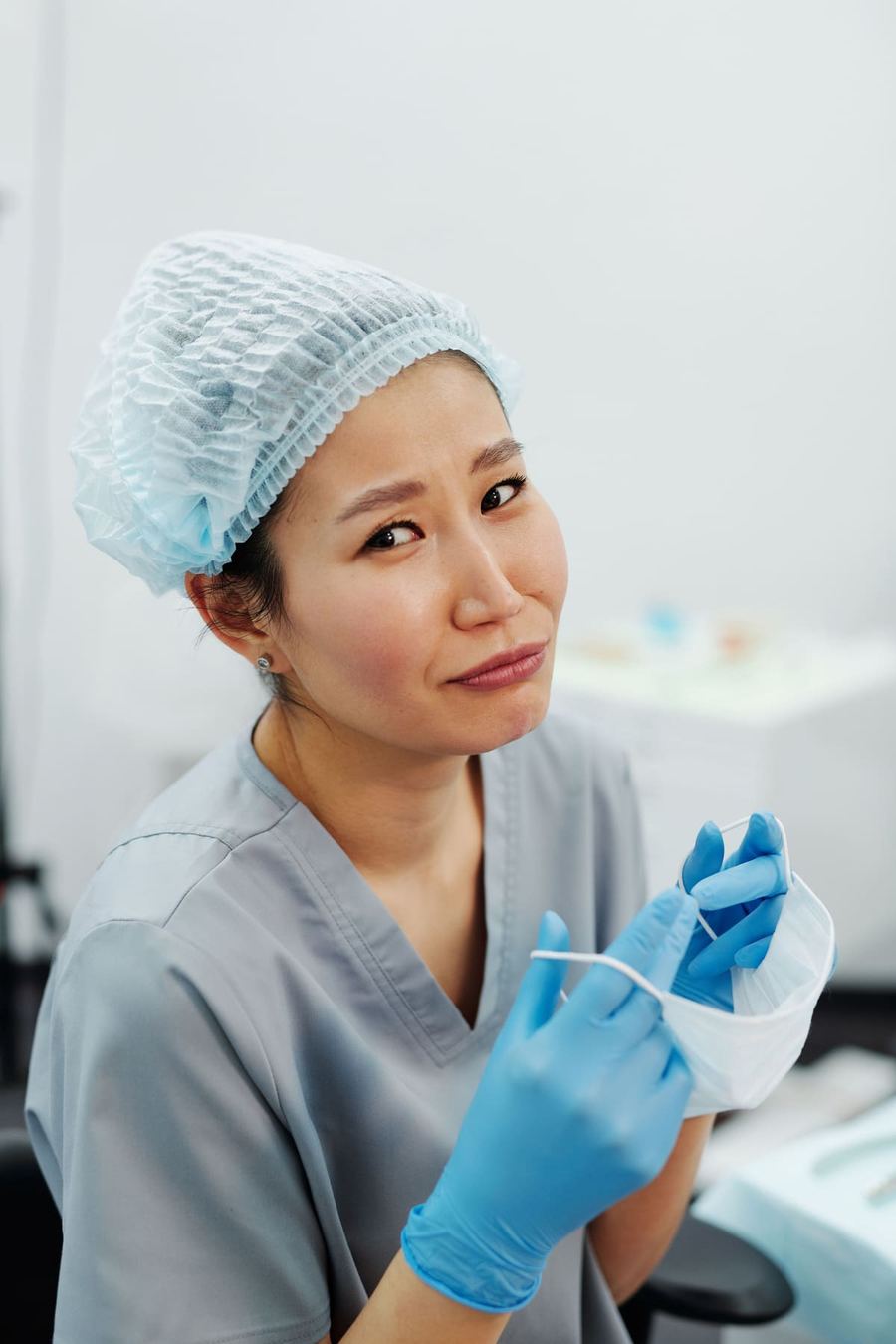 A dental assistant in a surgical cap and blue gloves holding a mask at a treatment chair.
