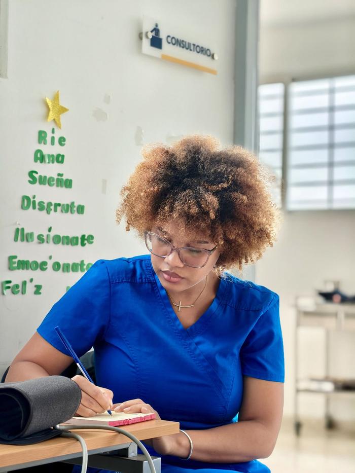 A medical assistant in blue scrubs writing notes at a desk beside a blood pressure cuff.