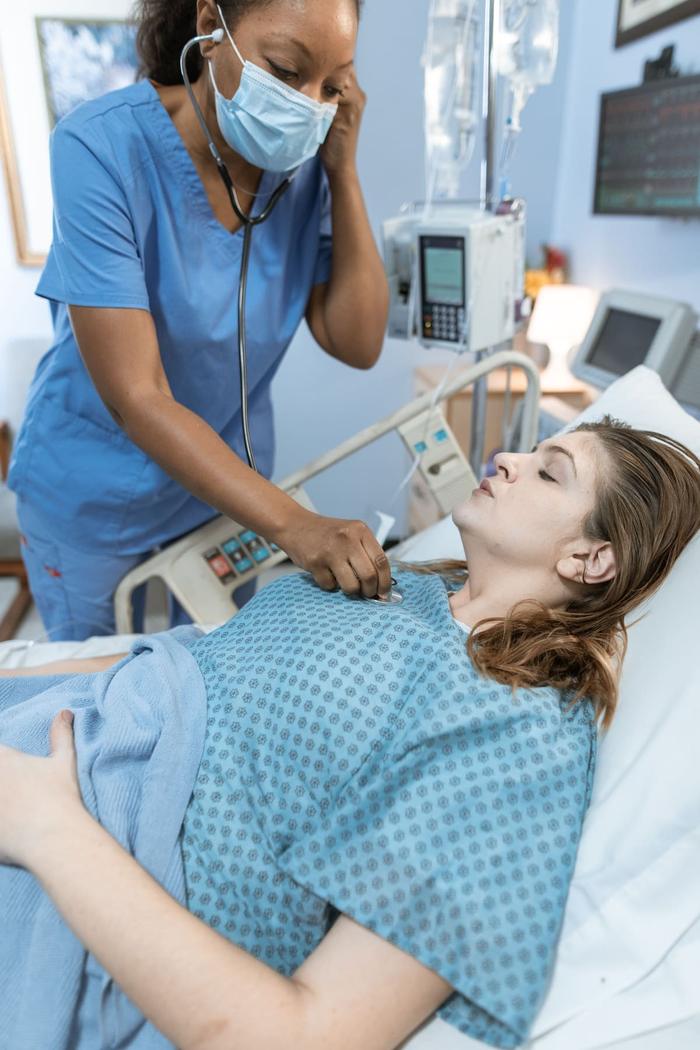 A clinician in blue scrubs using a stethoscope on a patient in a hospital bed beside an IV pump.