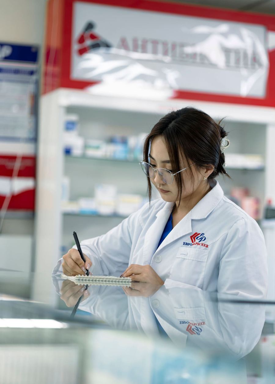 A pharmacy worker in a white coat filling a blister pack behind a pharmacy counter.