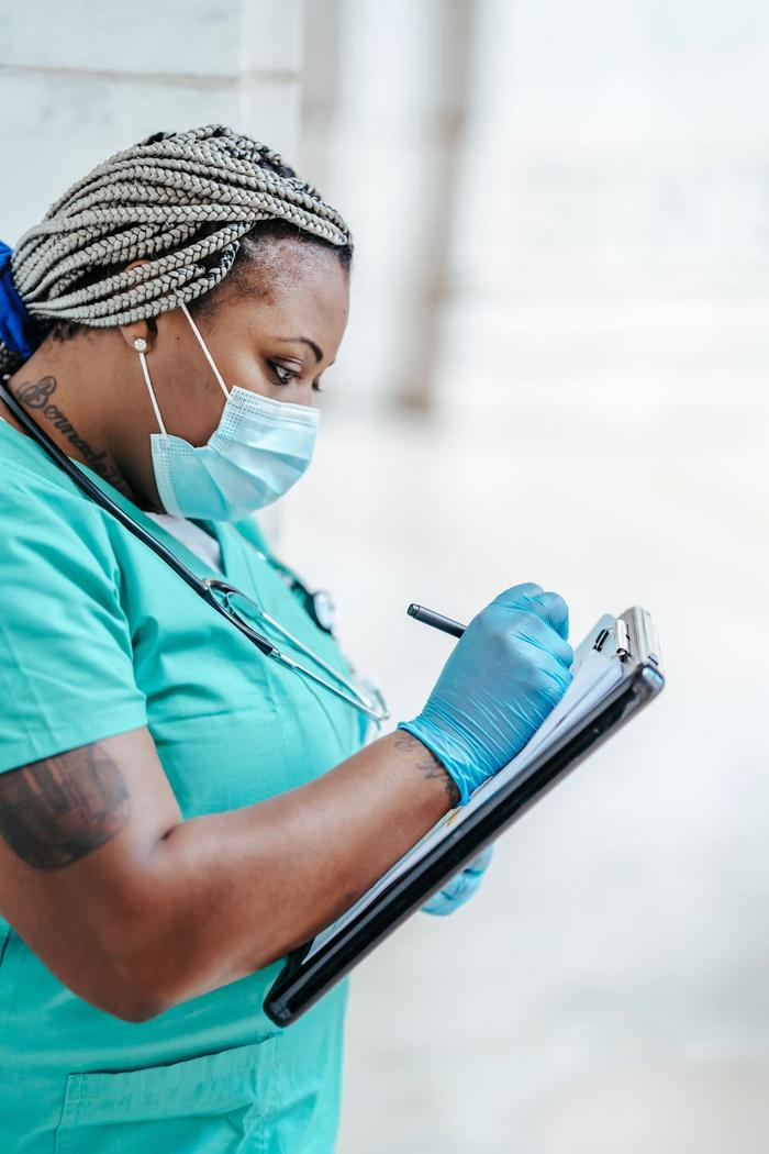 A healthcare worker in teal scrubs wearing a mask and gloves, writing on a clipboard.
