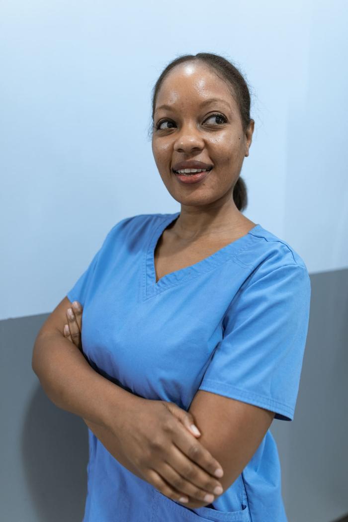 A healthcare worker in blue scrubs standing with arms folded, smiling warmly.
