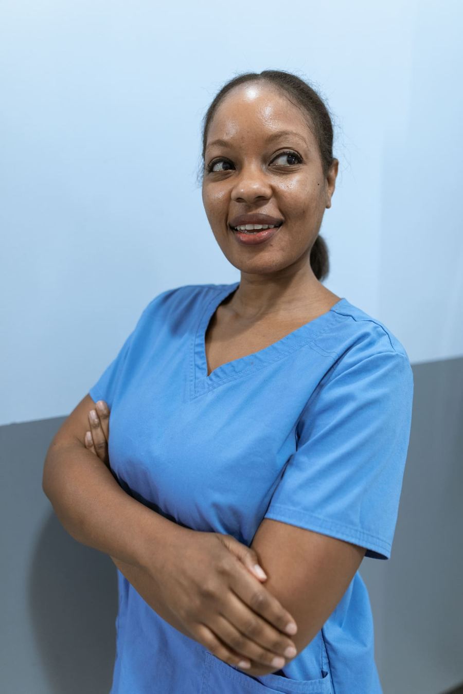 A healthcare worker in blue scrubs standing with arms folded, smiling warmly.