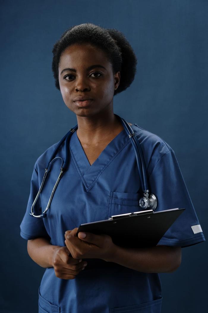 A nurse in blue scrubs holding a clipboard, a stethoscope draped around their shoulders.