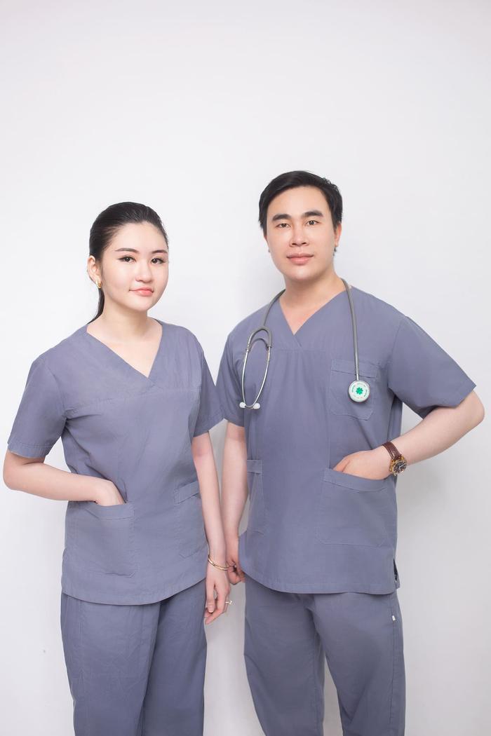 Two healthcare workers in grey surgical scrubs standing against a white wall.